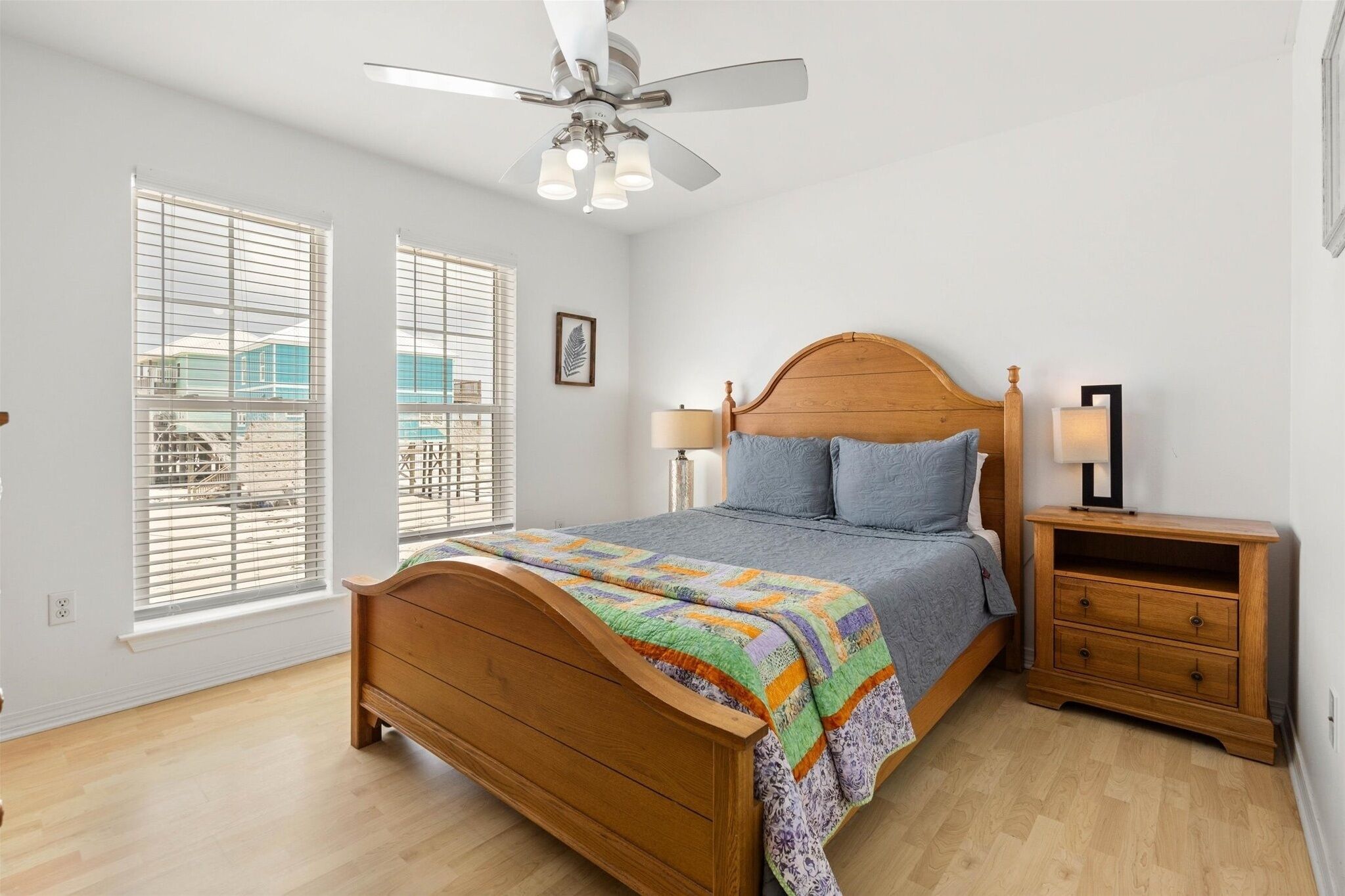 A bedroom with a wooden bed, two bedside tables, a ceiling fan, and large windows with blinds.