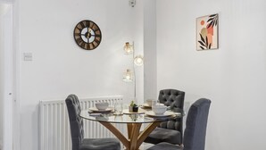 Un coin repas comprenant une table ronde en verre, des chaises à revêtement gris, une horloge murale, des luminaires suspendus et un tableau abstrait encadré.