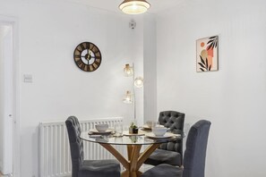 Un coin repas comprenant une table ronde en verre, des chaises à revêtement gris, une horloge murale, des luminaires suspendus et un tableau abstrait encadré.