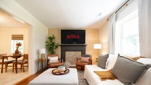 Una sala de estar con chimenea, un sofá, una mesa de comedor y un televisor que muestra 'NETFLIX'.