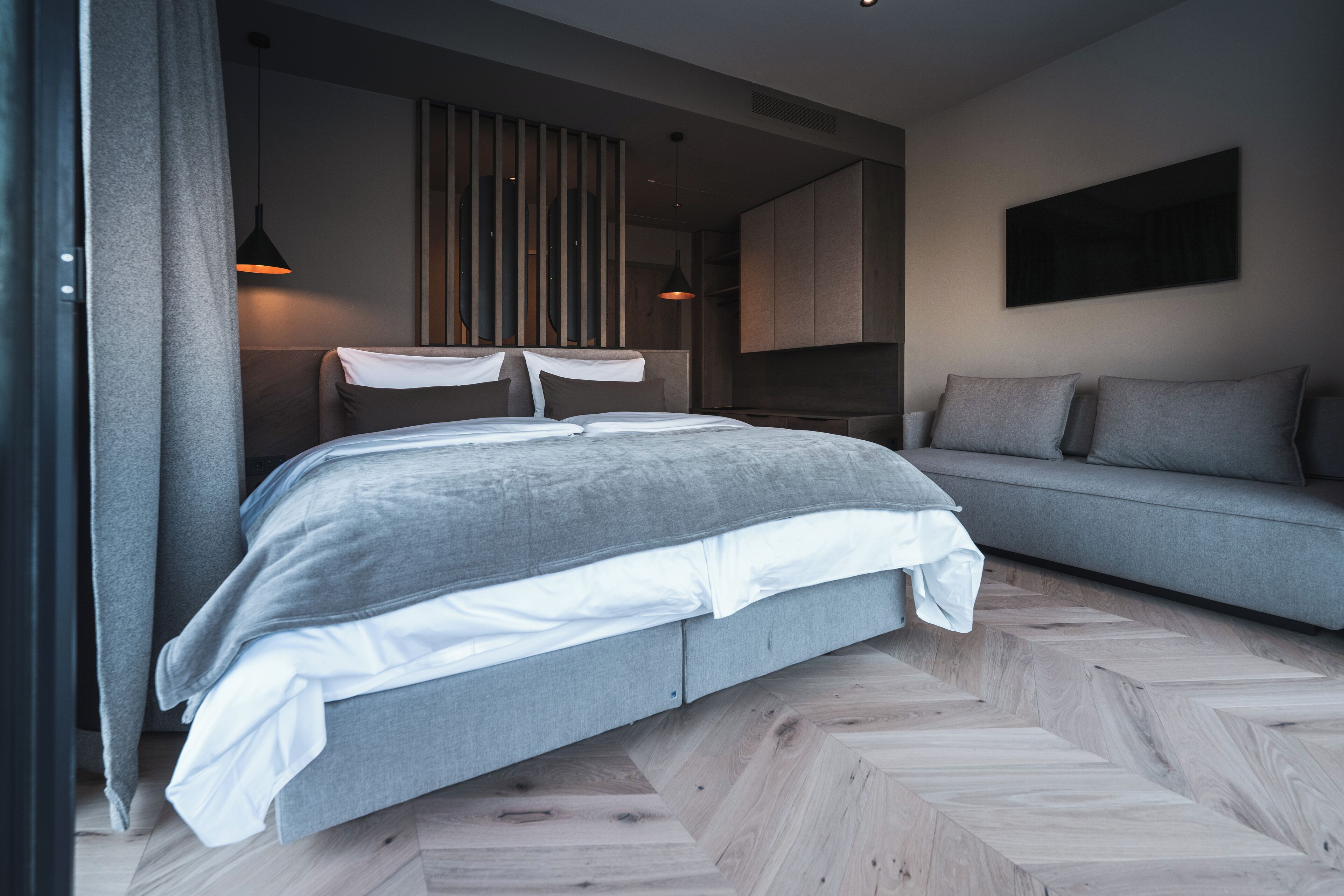 A modern bedroom with a bed, grey sofa, and a herringbone-patterned floor.