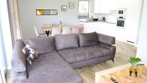 Ein modernes Wohnzimmer mit einer grauen Eckcouch, einem Essbereich mit Tisch und Stühlen sowie einer Küche mit weißen Schränken und Geräten.
