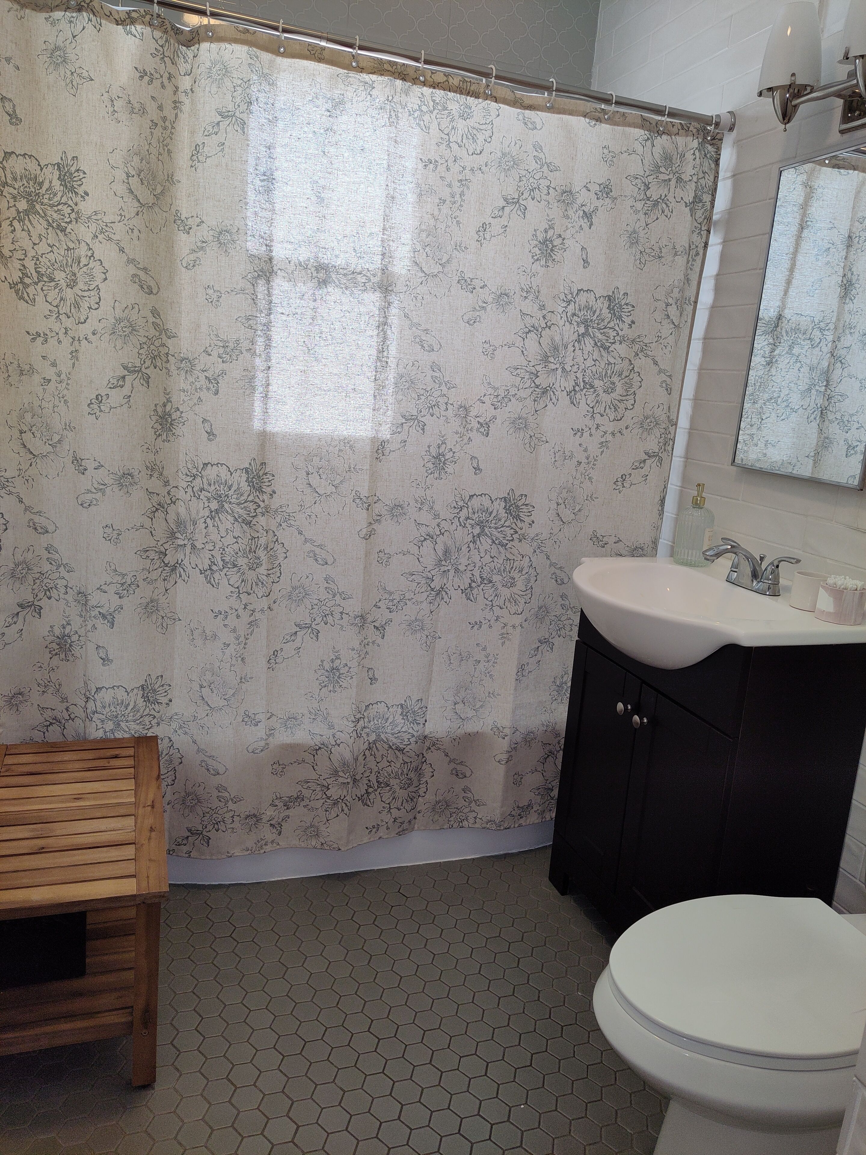 Un baño con cortina de ducha estampada con flores, un banco de madera, un lavamanos con espejo y un inodoro.