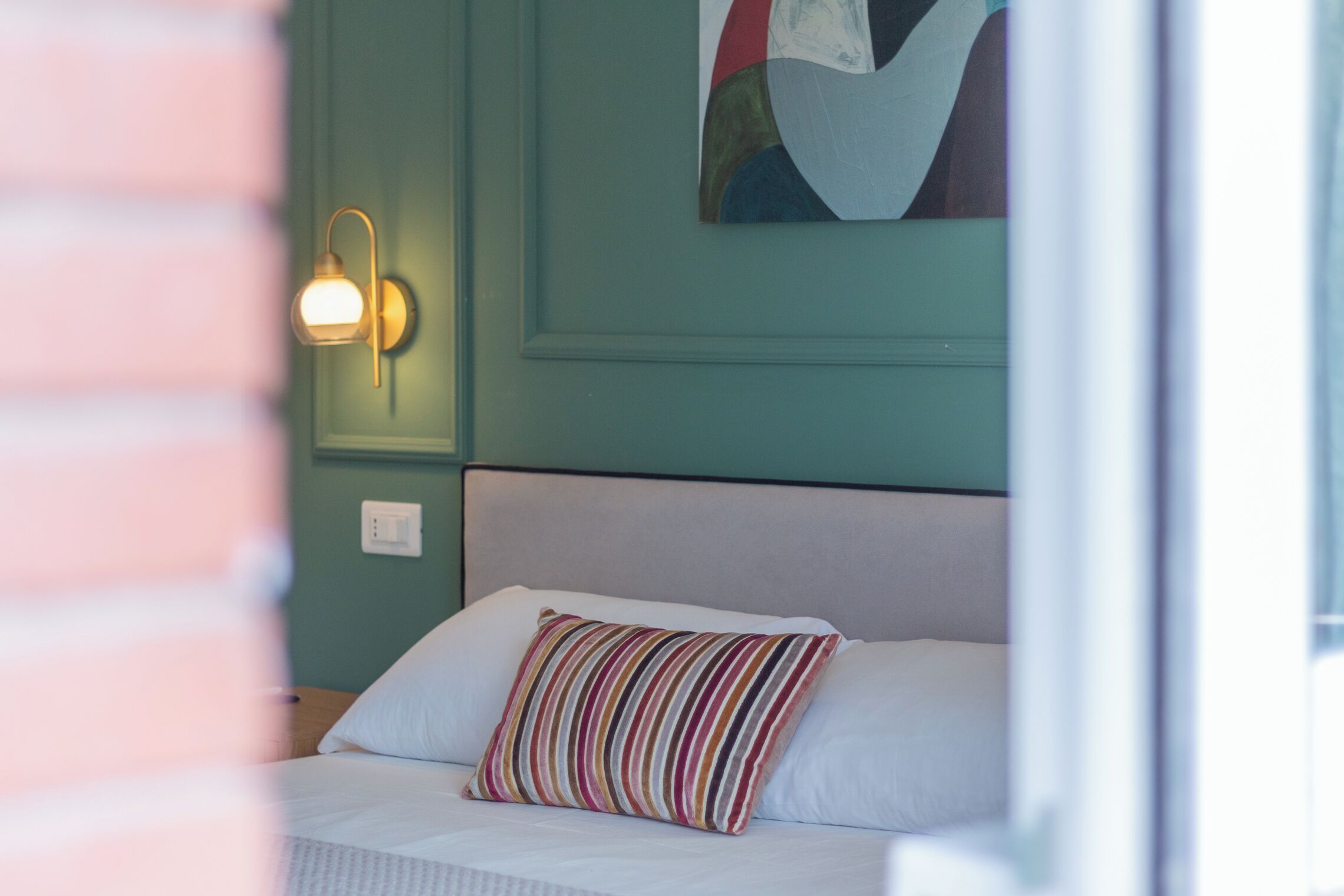 Un lit avec un oreiller à rayures, une lampe fixée au mur et une vue partielle d’un tableau.