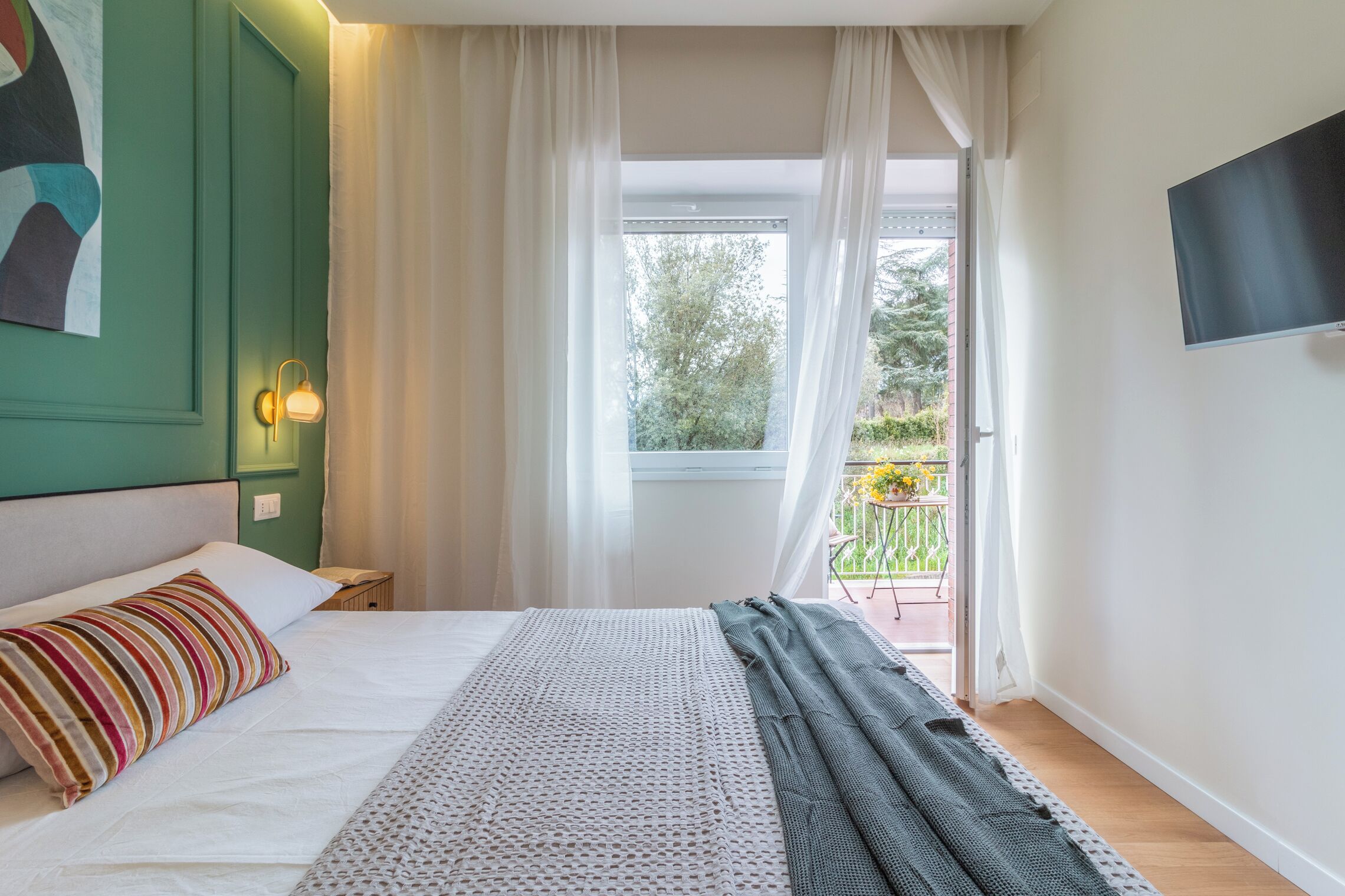 Un dormitorio con cama, mesita de noche, televisor montado en la pared y ventana con cortinas.