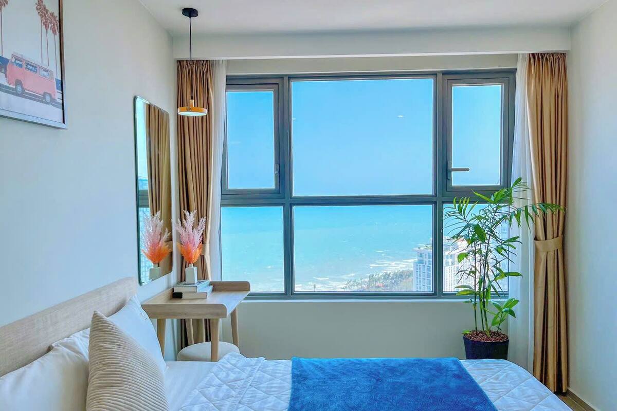 Habitación de hotel con un ventanal grande, una cama con manta azul, un escritorio y una planta en maceta.