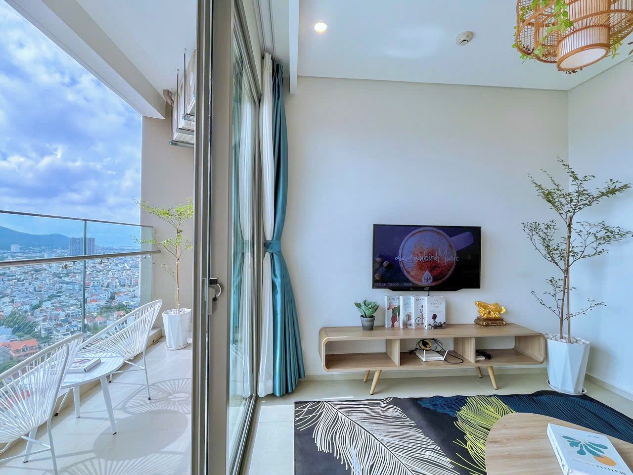 Una sala moderna con un televisor grande, un balcón con sillas blancas y vistas a la ciudad.