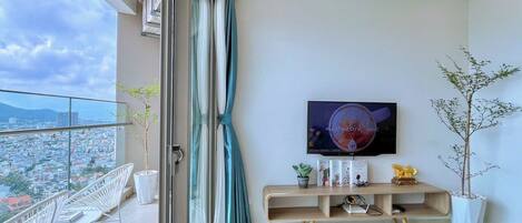 Una sala moderna con un televisor grande, un balcón con sillas blancas y vistas a la ciudad.