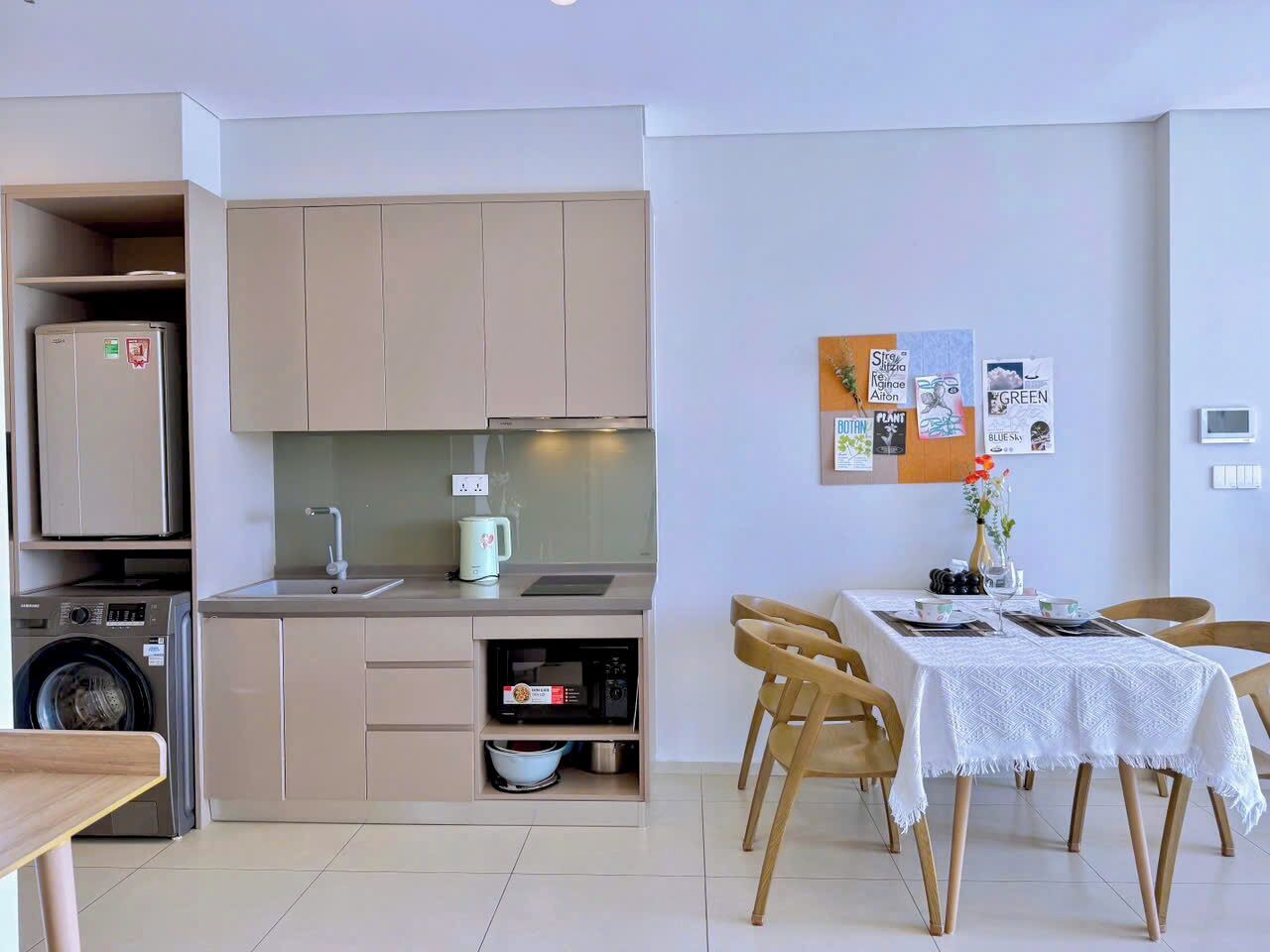 A compact kitchen with a washing machine, a microwave, and a sink. There is a dining area with a table and chairs, and a small plant on the wall.