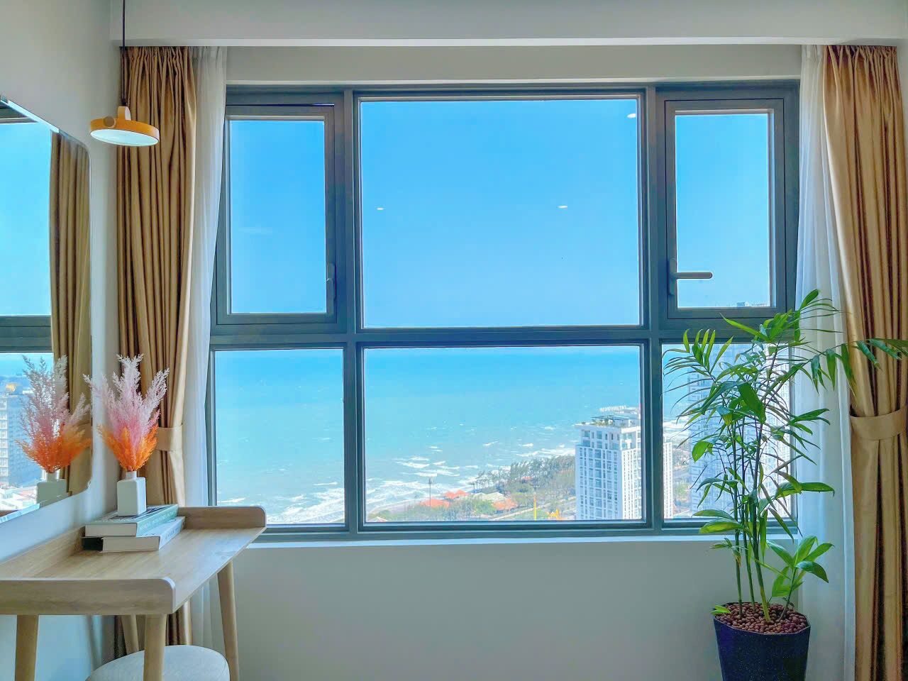 A room with a large window offering a view of the sea and buildings. There is a desk with a lamp, a potted plant, and decorative items.