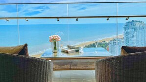 Un balcón con vistas a la playa y al océano, muebles de mimbre, una mesa de vidrio y un jarrón con flores.