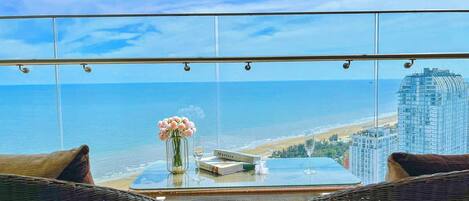 A balcony with a view of the beach and ocean, wicker furniture, a glass table, and a vase with flowers.