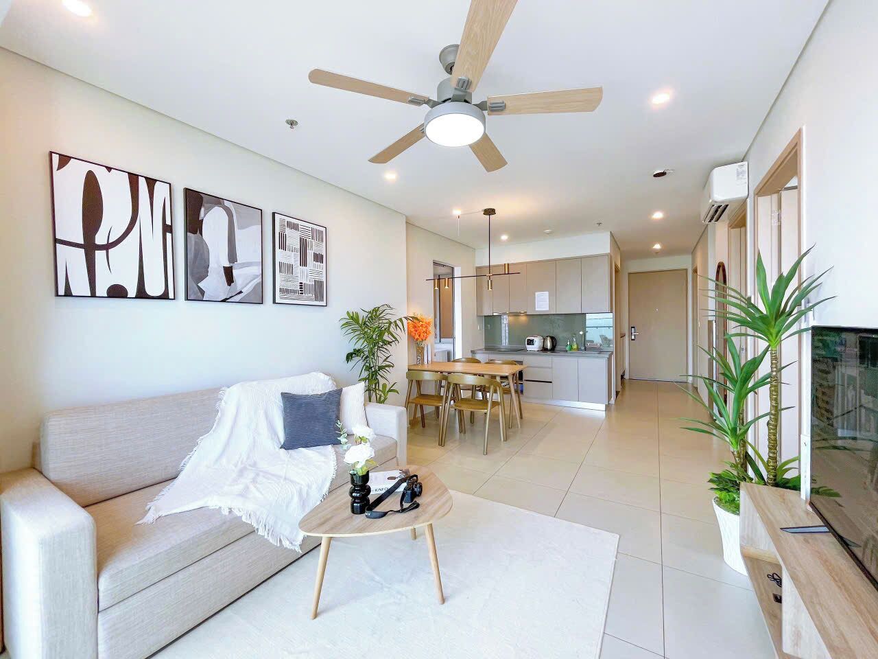 A modern living room with a sofa, coffee table, and artwork on the wall. There is a dining area with a table and chairs, and a kitchen in the background.