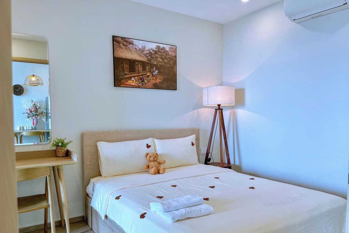 Una cama bien hecha con un osito de peluche, una lámpara de noche y un cuadro en la pared.