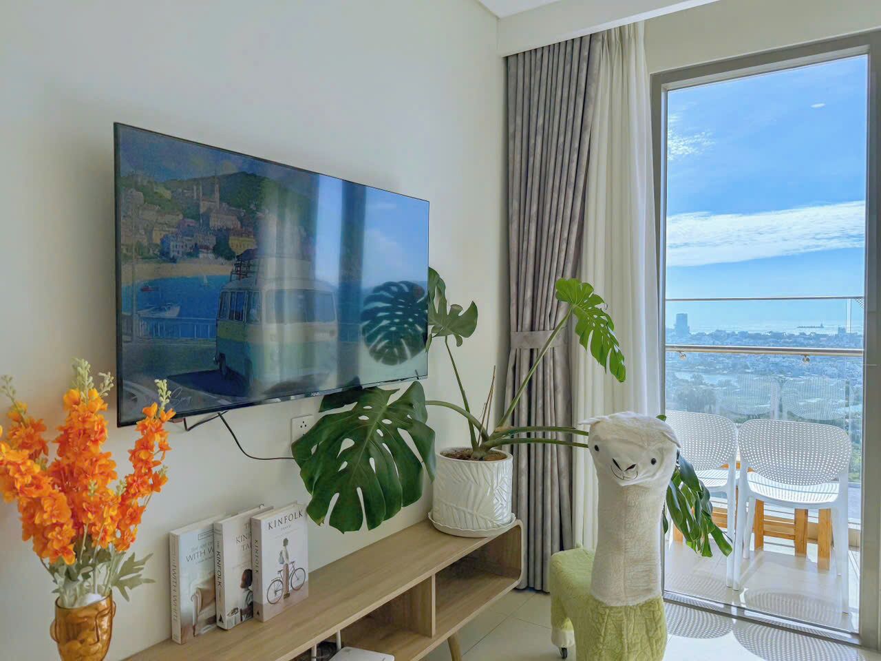 Un salon moderne avec un grand téléviseur à écran plat fixé au mur, un monstera en pot, une sculpture décorative blanche et une vue sur la ville grâce à de grandes fenêtres.
