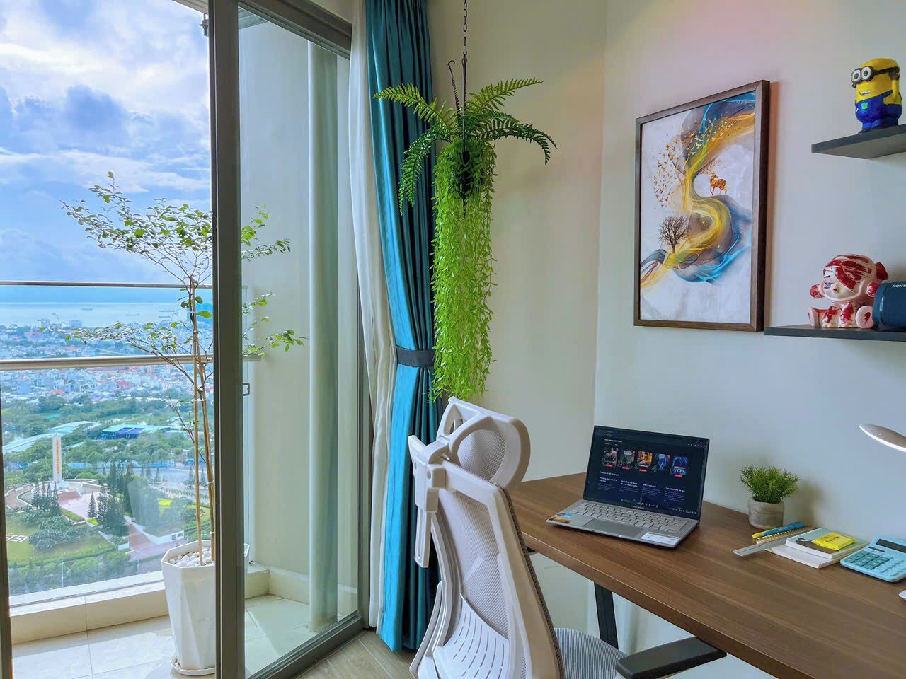 Une pièce moderne avec un ordinateur portable posé sur un bureau en bois, une plante suspendue, une chaise et une vue sur le paysage urbain à travers une grande fenêtre.