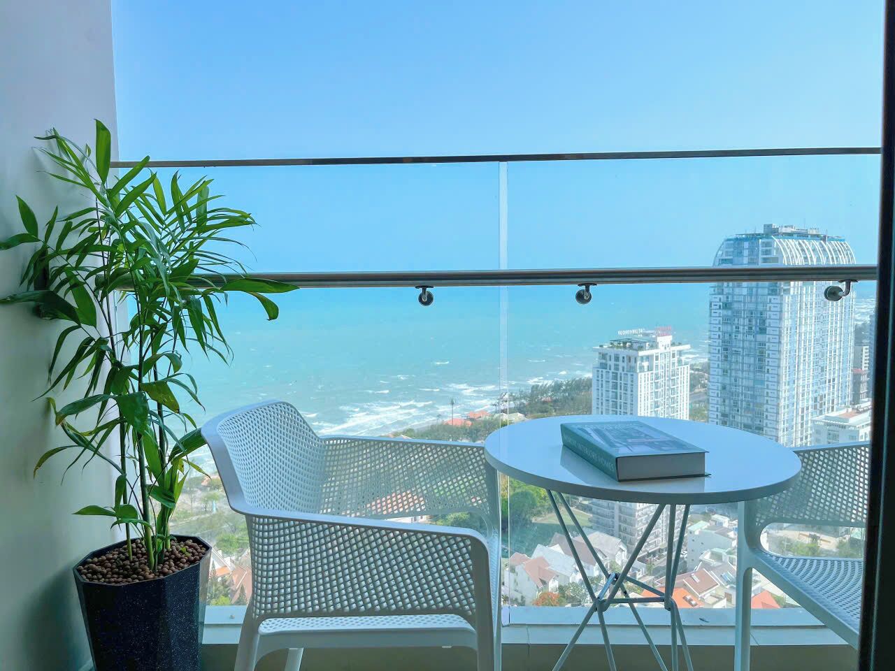 A balcony with a potted plant, two wicker chairs, and a small table with a book. A cityscape and ocean are visible through the window.