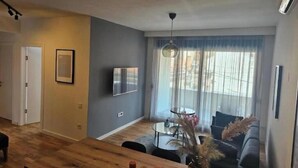 Een moderne woonkamer met een houten vloer, een ronde glazen tafel, een grijze bank en een groot raam met transparante gordijnen.