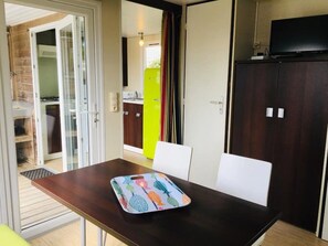 Un coin repas comprenant une table, des chaises et un set de table coloré. Une porte-fenêtre coulissante donne accès à une cuisine équipée d’un réfrigérateur et de placards.