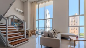 A modern living room with a staircase, a sofa, a dining table, and large windows offering a city view.