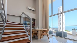 A modern interior with a wooden staircase, a dining area, and large windows offering a view of the sea and high-rise buildings.