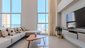 A modern living room with a white sofa, a coffee table, a TV, and large windows offering a city view.