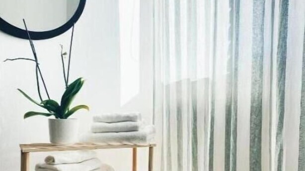 Una habitación luminosa con un estante de madera que sostiene toallas y una planta en maceta, cerca de un ventanal con cortinas de encaje.