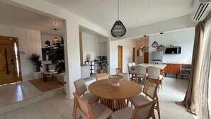 A spacious living area with a round wooden dining table surrounded by chairs, a kitchen area with modern appliances, and a large window with curtains.