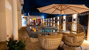 A poolside area with wicker chairs and an umbrella, a pool, and a building with a lit interior.