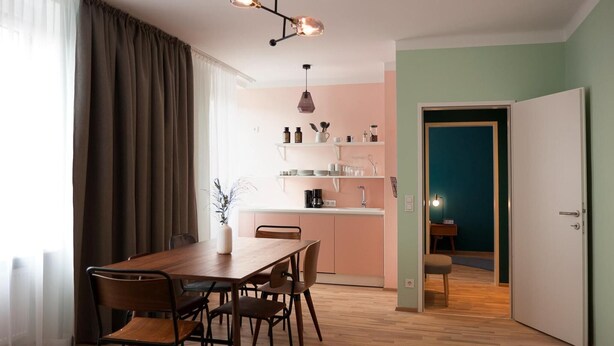 A dining area with a wooden table and chairs, a kitchen with shelves, and a door leading to another room. - Apartment in Linz near Danube Promenade (Linz)
