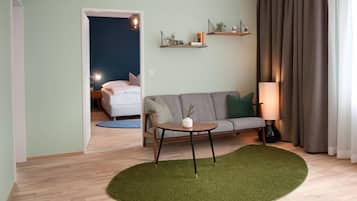 A living room with a sofa, a round table, and a lamp. There are two shelves on the wall with plants and books.