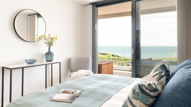 A bedroom with a bed, a bedside table with a vase and bowl, a chair, and a large window with a view of the sea. - 24 The Liner sits so close to the cove that the fresh sea breeze flows in through the windows. (Falmouth)