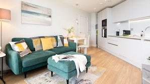 A modern living room with a teal sofa, a coffee table, and a dining area with a white table and chairs. - 24 The Liner sits so close to the cove that the fresh sea breeze flows in through the windows. (Falmouth)