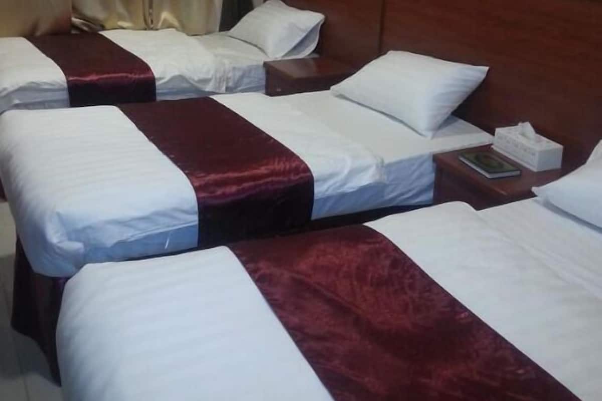 Habitación de hotel con tres camas, cada una con sábanas blancas y un detalle en rojo. Se observa un cabecero de madera, una mesita de noche con un libro y pañuelos, además de una pared con un estampado.