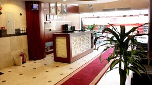 Een hotel lobby met een receptiebalie, een brandblusser, een plant en uitzicht op een winkel door het raam.