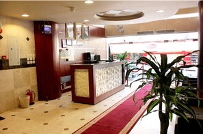 Un vestíbulo de hotel con un mostrador de recepción, un extintor, una planta y vista a una tienda a través del ventanal.