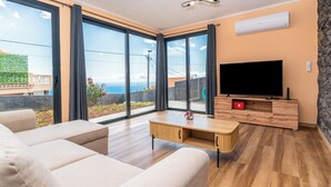 A modern living room with a large TV, wooden furniture, and a view of the sea.