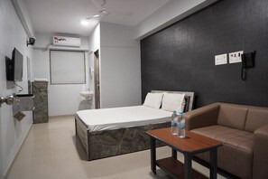 Een hotelkamer met een bed, een tafel met een fles water, een sofa, een televisie, een spiegel, een wastafel en een airconditioning.