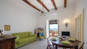 Ein Zimmer mit einem grünen Sofa, einem Esstisch mit Stühlen, einem gemusterten Boden und Blick durch eine offene Tür.