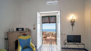 Ein Zimmer mit Kamin, einem Sessel, einem Fernseher auf einem Schrank und Blick auf das Meer durch eine offene Tür.