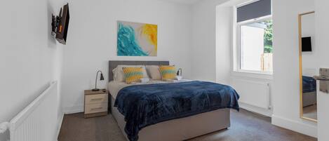 A bedroom with a bed, bedside table, lamp, artwork, window, and a mirror.