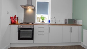 A modern kitchen with white cabinets, a stainless steel range hood, a built-in oven, a microwave, and a window with a view of a backyard. - Arran Cottage Spacious 4-Bedroom House (Coatbridge)