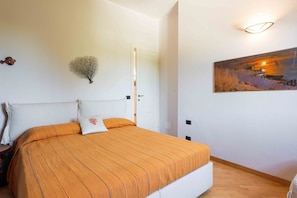A bedroom with a bed, white walls, a door, a painting, and a wall-mounted light fixture.