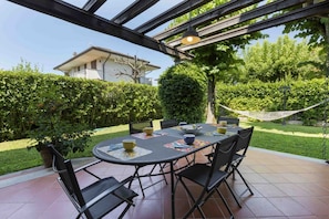 Une terrasse extérieure avec une table ronde et des chaises, entourée de verdure et agrémentée d’un hamac.