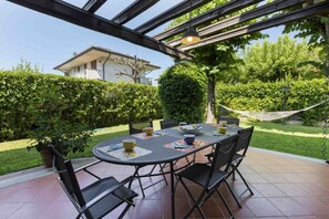 Een terras buiten met een ronde tafel en stoelen, omgeven door groen en een hangmat.