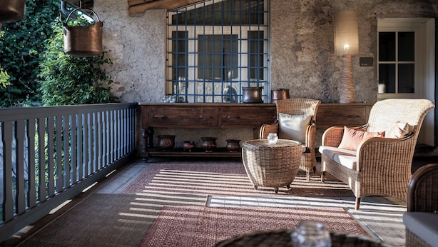 A rustic patio with wicker furniture, a wooden bench, and a view of greenery through a barred window.