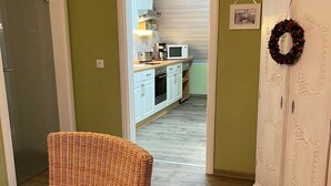 A room with a wicker chair, a white cabinet with a wreath, and a kitchen in the background. - To complete (Burgbrohl)