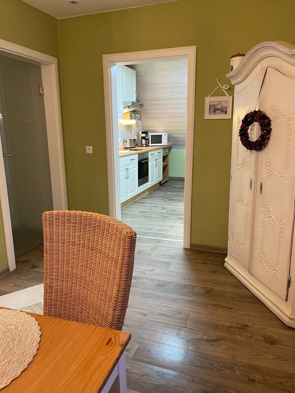 A room with a wicker chair, a white cabinet with a wreath, and a kitchen in the background. - To complete (Burgbrohl)