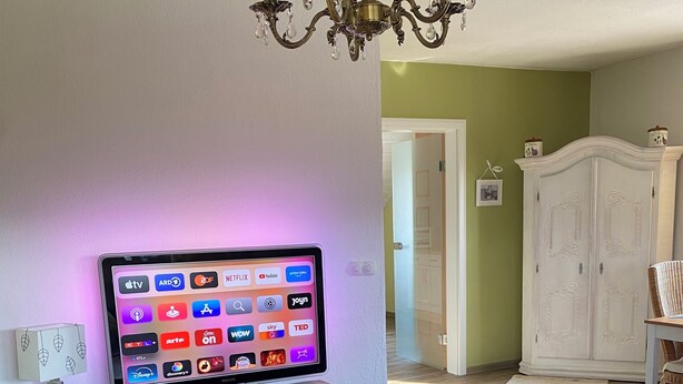 A living room with a flat-screen TV mounted on a wooden cabinet, a chandelier, and a white wardrobe in the background. - To complete (Burgbrohl)