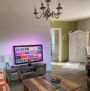 A living room with a flat-screen TV mounted on a wooden cabinet, a chandelier, and a white wardrobe in the background. - To complete (Burgbrohl)